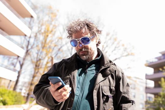 Mature man with grey hair in blue sunglasses looking at his smartphone while standing in the street. High quality photo