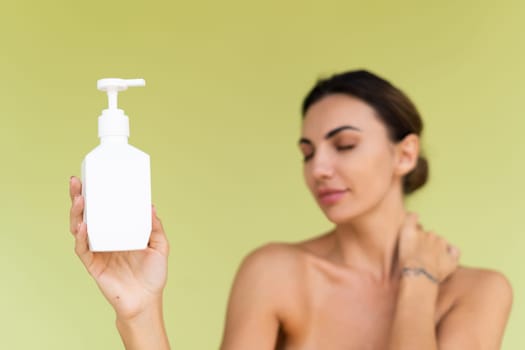 Beauty portrait of young topless woman with bare shoulders on green background with perfect skin and natural makeup holding bottle of shampoo, body lotion