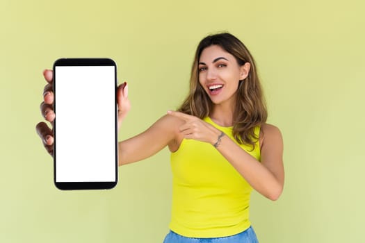 Young woman in casual wear isolated on green background holds a phone with a blank white screen