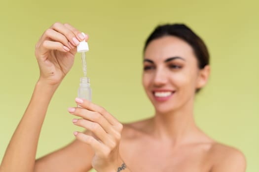 Beauty portrait of young topless woman with bare shoulders on green background with perfect skin and natural makeup holds serum for youth and skin hydration