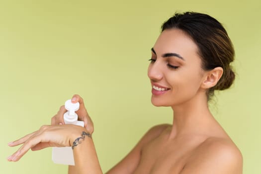 Beauty portrait of young topless woman with bare shoulders on green background with perfect skin and natural makeup holding bottle of shampoo, body lotion