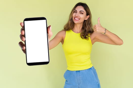 Young woman in casual wear isolated on green background holds a phone with a blank white screen