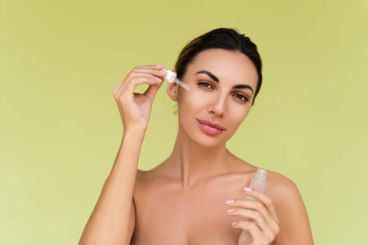 Beauty portrait of young topless woman with bare shoulders on green background with perfect skin and natural makeup holds serum for youth and skin hydration