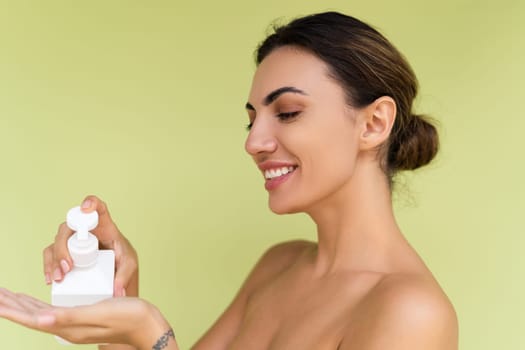 Beauty portrait of young topless woman with bare shoulders on green background with perfect skin and natural makeup holding bottle of shampoo, body lotion