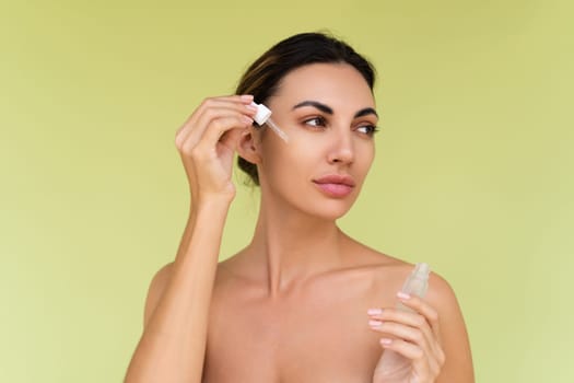 Beauty portrait of young topless woman with bare shoulders on green background with perfect skin and natural makeup holds serum for youth and skin hydration