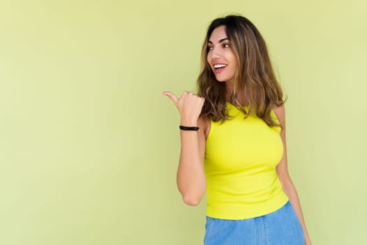 Young brunette in casual wear isolated on green background happy look to camera smile excited point thumb finger to left