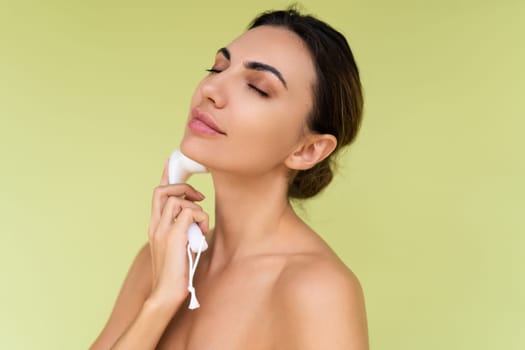 Beauty portrait of young topless woman with bare shoulders on green background with perfect skin and natural makeup holds face cleansing brush