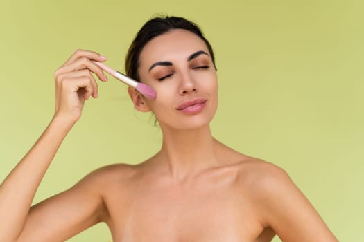 Beauty portrait of young topless woman with bare shoulders on green background with perfect skin and natural makeup holds blush brush