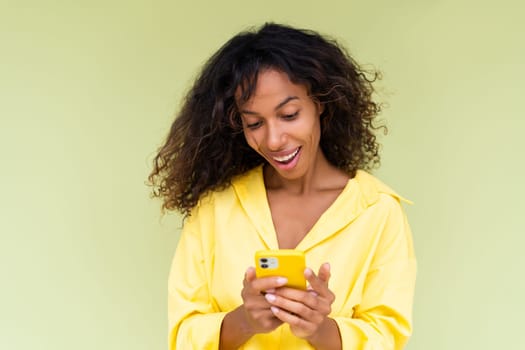 Beautiful african american woman in casual shirt on green background holds mobile phone with a smile