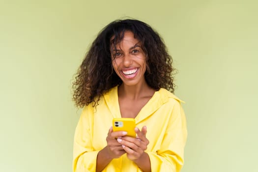 Beautiful african american woman in casual shirt on green background holds mobile phone with a smile