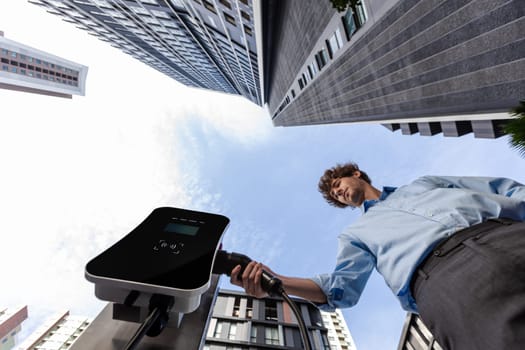 Fisheye view suit-clad progressive businessman at charging station for electric vehicle at modern city with the residential apartment condo building in the background. Idea for eco-friendly car.