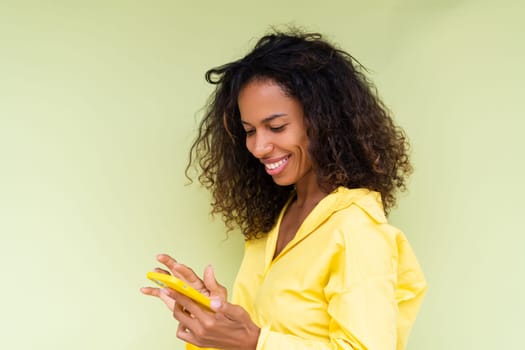 Beautiful african american woman in casual shirt on green background holds mobile phone with a smile