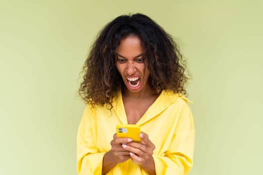 Beautiful african american woman in casual shirt on green background holds a phone look at screen with anger