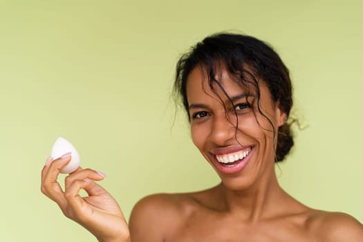 Beauty portrait of young topless african american woman with bare shoulders on green background with makeup foundation sponge