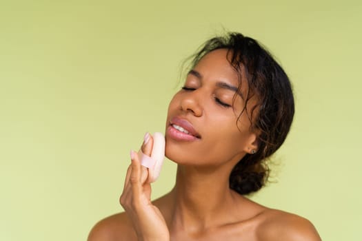 Beauty portrait of young topless african american woman with bare shoulders on green background with cosmetic powder puff velour makeup foundation blender sponge applicator