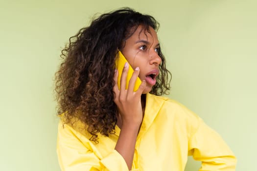 Beautiful african american woman in casual shirt on green background talk on mobile phone shocked confused