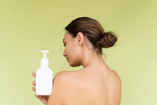 Beauty portrait of young topless woman with bare shoulders on green background with perfect skin and natural makeup holding bottle of shampoo, body lotion
