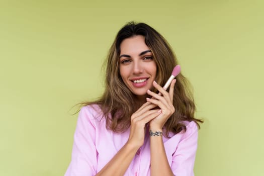 Young woman in casual wear isolated on green background playful positive with blush brush, natural daily makeup