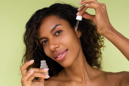Beauty portrait of young topless african american woman with bare shoulders on green background with perfect skin holds serum for youth and skin hydration