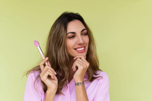 Young woman in casual wear isolated on green background playful positive with blush brush, natural daily makeup