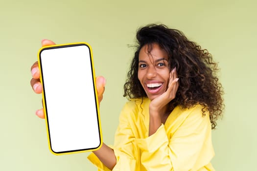 Beautiful african american woman in casual shirt on green background holds a phone with a blank white screen