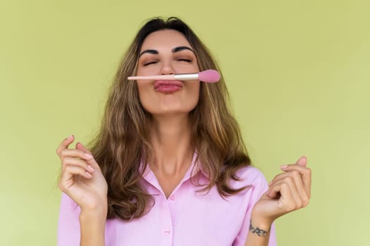 Young woman in casual wear isolated on green background playful positive with blush brush, natural daily makeup