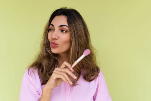 Young woman in casual wear isolated on green background playful positive with blush brush, natural daily makeup