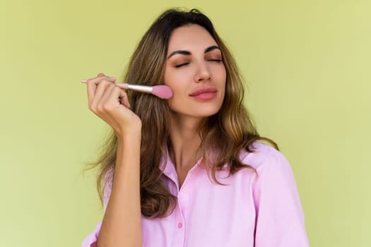 Young woman in casual wear isolated on green background playful positive with blush brush, natural daily makeup