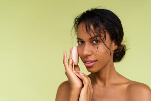 Beauty portrait of young topless african american woman with bare shoulders on green background with cosmetic powder puff velour makeup foundation blender sponge applicator