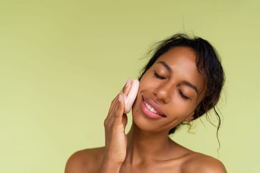 Beauty portrait of young topless african american woman with bare shoulders on green background with cosmetic powder puff velour makeup foundation blender sponge applicator