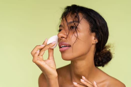 Beauty portrait of young topless african american woman with bare shoulders on green background with makeup foundation sponge