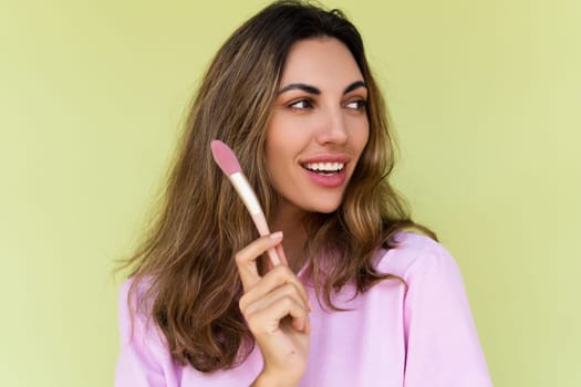 Young woman in casual wear isolated on green background playful positive with blush brush, natural daily makeup