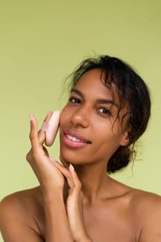Beauty portrait of young topless african american woman with bare shoulders on green background with cosmetic powder puff velour makeup foundation blender sponge applicator