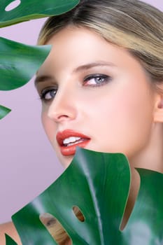 Closeup woman with perfect clean skin and alluring flawless natural soft facial makeup holding green leave monstera. Natural skincare treatment beauty or spa concept in pink isolated background.