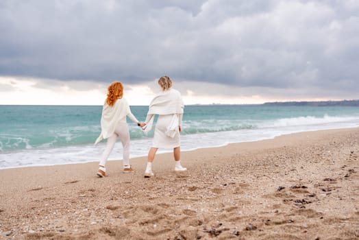 Women sea walk friendship spring. Two girlfriends, redhead and blonde, middle-aged walk along the sandy beach of the sea, dressed in white clothes. Against the backdrop of a cloudy sky and the winter sea. Weekend concept