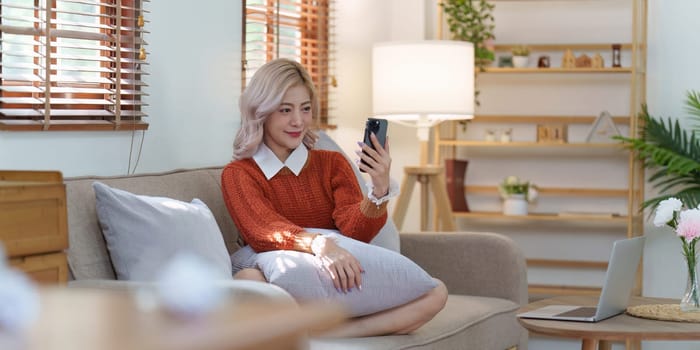 Beautiful Asian Woman relaxing at home and using mobile phone sitting on cozy sofa.