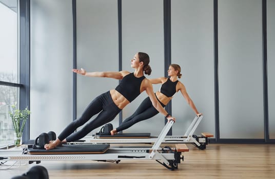 Standing on gym equipment and doing stretches. Two women in sportive wear and with slim bodies have fitness yoga day indoors together.