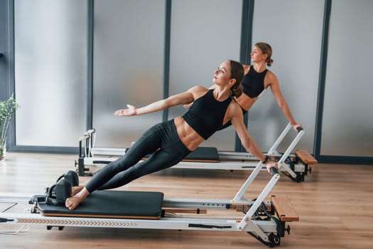 Standing on gym equipment and doing stretches. Two women in sportive wear and with slim bodies have fitness yoga day indoors together.