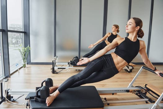 Standing on gym equipment and doing stretches. Two women in sportive wear and with slim bodies have fitness yoga day indoors together.