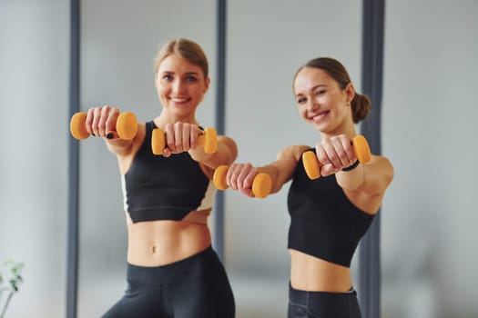 Uses dumbbells. Two women in sportive wear and with slim bodies have fitness yoga day indoors together.