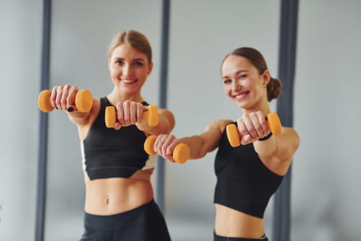 Uses dumbbells. Two women in sportive wear and with slim bodies have fitness yoga day indoors together.