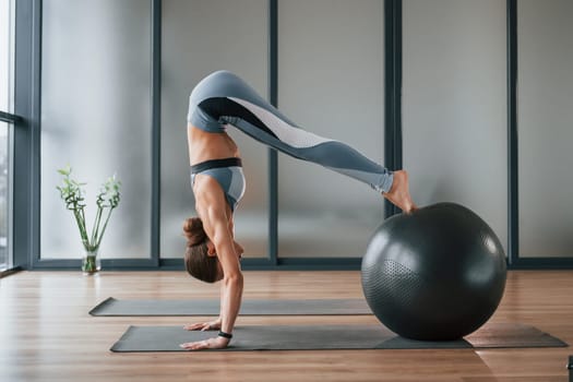 Uses fitness ball. Young woman in sportive wear and with slim body have yoga day indoors.