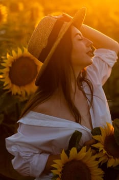 A girl in a hat on a beautiful field of sunflowers against the sky in the evening light of a summer sunset. Sunbeams through the flower field. Natural background