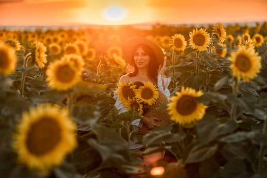 A girl in a hat on a beautiful field of sunflowers against the sky in the evening light of a summer sunset. Sunbeams through the flower field. Natural background