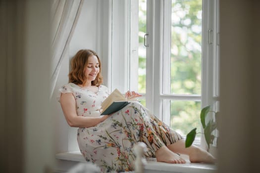 A middle-aged woman in a cream dress sits mysteriously and looks out the window on the windowsill. Green trees outside