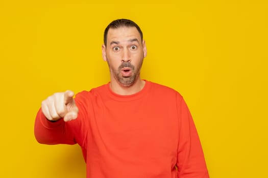 Bearded hispanic man wearing a red sweater pointing towards the camera with a surprised expression and open mouth, isolated over yellow background