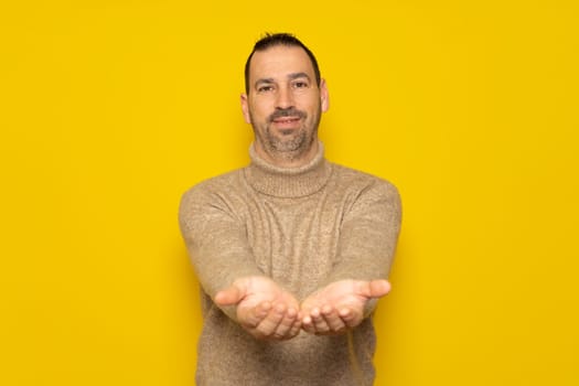 Bearded hispanic handsome man dressed in a beige turtleneck offering something in his hands, has a friendly and receptive attitude. Isolated on yellow background.