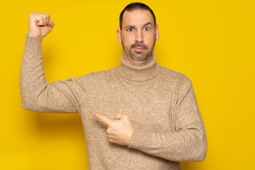 Portrait bearded latino man pointing finger at arm muscle, discount, looking at camera with satisfied face. Indoor studio shot isolated on yellow background