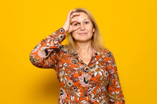 Caucasian blonde woman in patterned dress doing ok gesture surprised with shocked face, looking into eyes through fingers. Incredulous expression