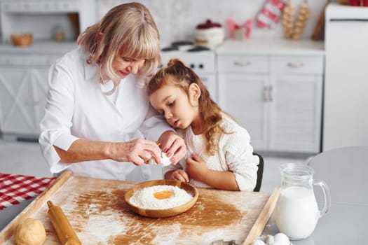 Uses eggs. Senior grandmother with her little granddaughter cooks sweets for Christmas on the kitchen.